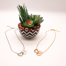Load image into Gallery viewer, Gold tone and silver tone necklaces with open heart charms intertwined with open pet paw charms. Displayed on white background with succulent plant as accent.