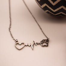 Load image into Gallery viewer, Silver necklace with open heart, heartbeat rhythm, and open pet paw. Closeup on white background with partial image of pot in background.