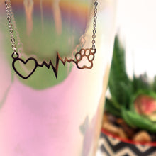 Load image into Gallery viewer, Silver necklace with open heart, heartbeat rhythm, and open pet paw. Displayed hanging on side of irridescent cup with succulent in background.