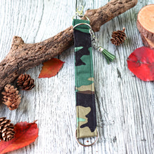 Load image into Gallery viewer, Closeup of the green camo Charmable Wristlet keychain. Displayed on a wood with a branch, orange leaves and pine cones.