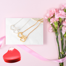 Load image into Gallery viewer, Gold tone and silver tone necklaces with open heart charms intertwined with open pet paw charms. Displayed on white with pink background, pink flowers and red heart candy tin.