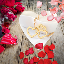 Load image into Gallery viewer, Gold tone necklace with open heart charms intertwined with open pet paw charms. Displayed hanging over edge of white bowl on background with wood, roses, and flower petals.