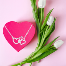 Load image into Gallery viewer, Silver tone necklace with open heart charms intertwined with open pet paw charms. Displayed laying on pink heart candy box with tulips beside it.