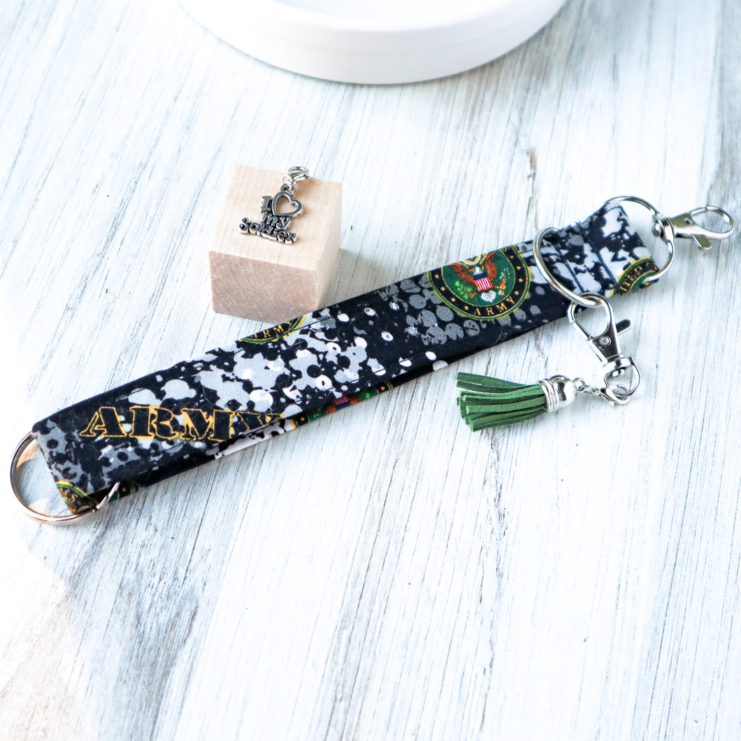 closeup picture of an Army Charmable Wristlet keychain on a white wood with a white bowl in the background and a charm on small light wood blocks. 