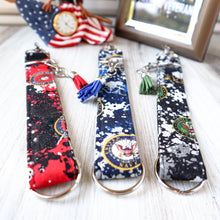 Load image into Gallery viewer, Closeup Picture of Marine, Navy and Army Charmable Wristlet keychain.  Displayed on a white board with a window in the background, an eagle clock and a picture thanking veterans. 
