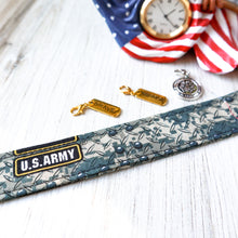 Load image into Gallery viewer, Closeup of an Army Digital pattern Charmable Wristlet keychain on white wood with an eagle clock and Brave, Warrior and Army Seal charms in the background. 
