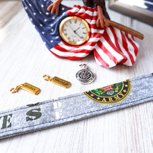 Load image into Gallery viewer, Closeup of an Army Motto pattern Charmable Wristlet keychain on white wood with an eagle clock and three charms in the background (Brave, Warrior, Army Seal). 
